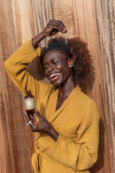 Eine lächelnde Frau mit lockigem Haar und einem gelben Bademantel trägt mit einer Pipette Haaröl auf ihre Kopfhaut auf, vor einem Holzhintergrund.