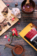 Aufnahme von oben von einer Packung „Love Choc Adaptogenic Cacao Elixir“, die diagonal auf einer dunklen Holzoberfläche platziert ist. Sie ist umgeben von Kakaopulver, einem Holzlöffel mit Pulver, einer kleinen Schale mit Pulver und Rosenblättern, Schokoladenstücken auf einem Schneidebrett, einer roten Rose und einem klaren Quarzkristall-Cluster.