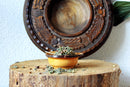 Eine kleine, gelb-braune Schale, gefüllt mit getrockneter Kräuterteemischung, die auf einem hölzernen Baumstumpf vor einem dekorativen geschnitzten Holzteller steht.