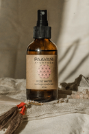Eine braune Sprühflasche "PAAVANI AYURVEDA ROSE WATER soothing aromatherapy" (4 fl. oz.) steht auf einem beigefarbenen, strukturierten Stoff, umgeben von gold-rosa Perlen und einem braunen Quastenanhänger, was eine entspannende Atmosphäre schafft.