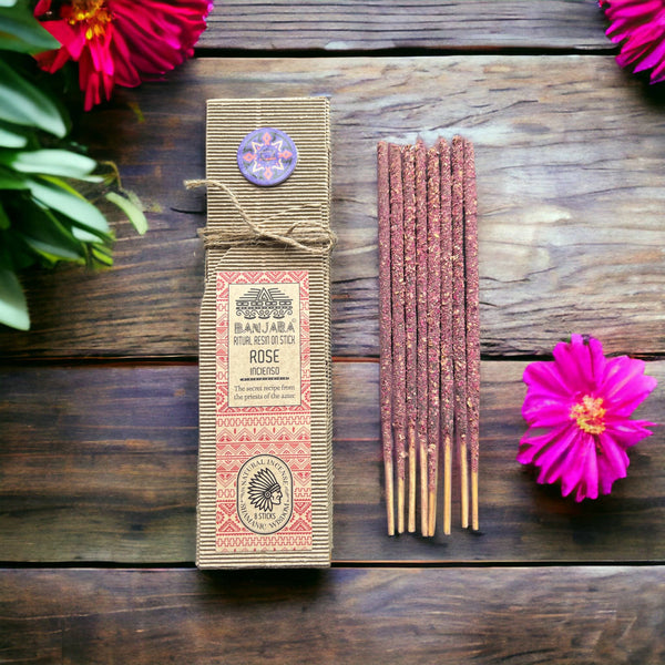 Eine Schachtel BANJARA Ritual Resin on Stick ROSE Incenso Räucherstäbchen neben einem Bündel pink-roter Räucherstäbchen, arrangiert auf dunklem Holz mit leuchtend pinken Blüten.