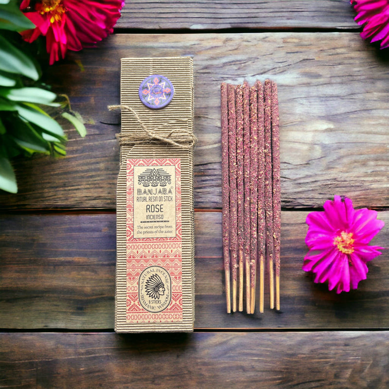 Eine Schachtel BANJARA Ritual Resin on Stick ROSE Incenso Räucherstäbchen neben einem Bündel pink-roter Räucherstäbchen, arrangiert auf dunklem Holz mit leuchtend pinken Blüten.