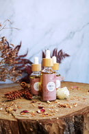 Drei kleine Flaschen mit Pipetten und goldener Flüssigkeit, die als "Freya" gekennzeichnet sind, stehen auf einem Baumstumpf, umgeben von getrockneten Kräutern und einer weißen Rose.