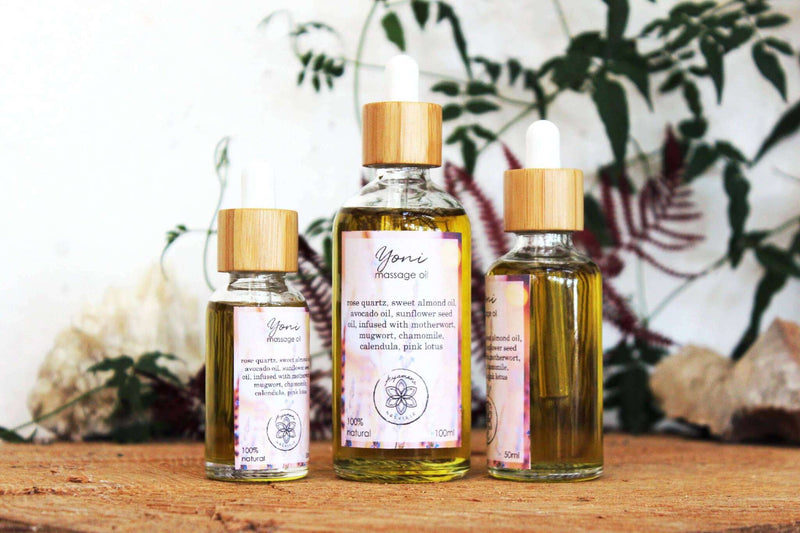 Drei Glasflaschen mit Pipette für "Yoni Massage Oil" mit Holzverschlüssen stehen aufrecht auf einer strukturierten Holzoberfläche, möglicherweise einem Baumstumpf. Die Flaschen variieren in der Größe, und eine zarte grüne Ranke sowie ein leuchtend roter Farn bilden natürliche Akzente im Hintergrund, alles vor einer unscharfen hellen Wand.