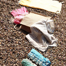 Verschiedene Hamamtücher in Pink, Beige, Gelb, Blau und Grün liegen zusammen mit einer braunen Jutetasche auf einem Bett aus kleinen, runden Kieselsteinen.