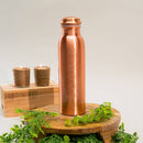 Eine hohe Kupferwasserflasche, graviert mit den sieben Chakren, präsentiert auf einem kleinen Holzständer mit zwei brennenden Kerzen und grünem Laub.