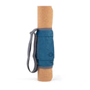 Eine zusammengerollte Kork-Yogamatte, die aufrecht steht. Sie ist mit einem grauen, verstellbaren Schultergurt und einer zentralen blau-grauen Tragetasche mit Reißverschlusstasche gesichert und vor einem weißen Hintergrund abgebildet.