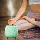 Ein dekorativer, abgerundeter Ultraschall-Aromadiffusor in weißer Lamellenoptik, der grün leuchtet und Dampf abgibt. Er steht auf dem Boden vor einer Person, die in einer Yoga-Pose meditiert.