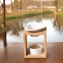 Eine Aromalampe aus Bambusholz mit einer herzförmigen Keramikschale, auf einem Holztisch vor einem unscharfen Hintergrund mit Naturlandschaft und einem Teich.