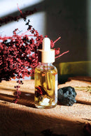 Eine einzelne Glasflasche mit Pipette und Holzverschluss für "Ritualöl" steht auf einer hellen Holzoberfläche, gefüllt mit goldener Flüssigkeit und sichtbaren Pflanzenbestandteilen. Daneben liegt ein rauer schwarzer Stein, und rote Trockenblütenzweige sind im Vorder- und Hintergrund zu sehen, während Sonnenlicht Schatten auf eine graue Wand wirft.