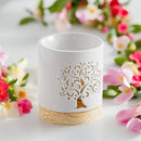 Eine weiße Keramik-Duftlampe oder Aromalampe mit einem ausgeschnittenen Lebensbaum-Motiv auf einem Holzsockel, umgeben von rosa und weißen Blumen.