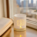 Eine beleuchtete, weiße Keramik-Duftlampe mit einem leuchtenden Lebensbaum-Ausschnitt, die auf einem Fensterbrett neben einem aufgeschlagenen Buch steht, mit einer Stadtlandschaft im Hintergrund.