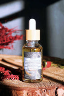 Eine Nahaufnahme einer Glasflasche mit Pipette und Holzverschluss für "Ritualöl", gefüllt mit goldener Flüssigkeit und infundierten Pflanzen. Sie steht auf einer Holzoberfläche mit sichtbarer Holzmaserung, während unscharfe rote Trockenblumen im Vorder- und Hintergrund eine natürliche und warme Atmosphäre schaffen.