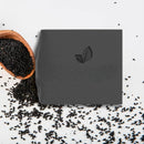 Quadratisches Stück schwarzer Seife mit Blatt-Logo, neben Schwarzkümmelsamen.