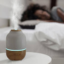 Aroma-Diffusor aus Holz und grau-weißer Keramik mit blauem Licht auf einem Nachttisch vor einer schlafenden Person.