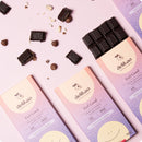 Zwei Packungen "delikao Feel Good" 70% Zartbitterschokolade mit Haselnusspraline, zusammen mit Schokoladenstücken und Haselnüssen auf einem rosa Hintergrund.