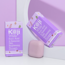 Lilafarbenes Bild mit zwei Schachteln Koji White Brightening Soap mit Kojisäure und Hyaluronsäure. Davor befindet sich ein rosa Seifenstück.