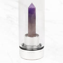 Nahaufnahme einer transparenten Wasserflasche mit einem spitz zulaufenden violetten Amethystkristall auf einem silbernen Sockel, der auf einer weißen Marmoroberfläche steht.