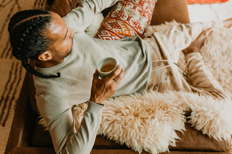 Ein Mann mit geflochtenen Haaren liegt entspannt auf einem braunen Sofa mit einer pelzigen Decke und hält eine Tasse Tee in der Hand, umgeben von einem gemusterten Kissen.