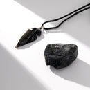 Ein Stück roher schwarzer Obsidian liegt neben einem passenden Obsidian-Pfeilspitzen-Anhänger an einer schwarzen Kordel, auf einer weißen Oberfläche mit starker gerichteter Beleuchtung fotografiert.