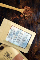 Zwei „Ayamanu Naturals“ Superfood-Pulverbeutel, darunter ein „Kaffee-Ersatz“, sind auf einer dunklen Holzoberfläche arrangiert. Ein Holzlöffel mit Pulver und verstreute Kakaobohnen vervollständigen die natürliche und organische Ästhetik.