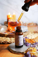 Eine Pipette tropft goldene Flüssigkeit in eine bernsteinfarbene Glasflasche mit der Aufschrift „Blue Lotus Tincture“ auf einem runden weiß-goldenen Untersetzer. Im Hintergrund sind verschwommen brennende Kerzen, ein Glas bernsteinfarbener Flüssigkeit mit einer Blume und getrocknete lila und orangefarbene Pflanzen auf einer gewebten Matte zu sehen.