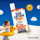Ein lächelndes Kind mit Enten-Schwimmhilfe und einer Flasche Harmonify Sonnenlotion Kids LSF 50+, betont "SEHR HOHER SCHUTZ" vor einem hellblauen und weißen Strandhintergrund.