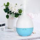 Ein weiß-blauer tropfenförmiger Aroma-Diffusor auf einer marmorierten Oberfläche neben einer Vase mit weißen Blumen auf einem Nachttisch.