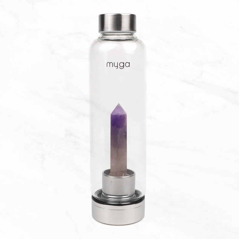  Eine transparente "myga" Wasserflasche mit einem spitz zulaufenden violetten Amethystkristall auf einem silbernen Sockel, stehend auf einer weißen Marmoroberfläche.