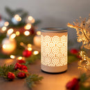 Eine beleuchtete, keramische Duftlampe/Wärmediffusor mit einem ausgeschnittenen Kreismuster in einer weihnachtlichen Umgebung mit Beeren und Lichterketten.