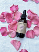 Eine braune Sprühflasche "PAAVANI AYURVEDA ROSE WATER soothing aromatherapy" (4 fl. oz.) liegt auf einer weißen Oberfläche, umgeben von verstreuten pinken Rosenblättern, was eine beruhigende Atmosphäre suggeriert.