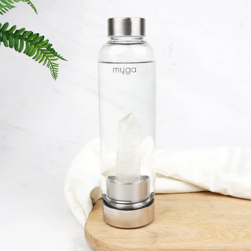 Eine klare Glaswasserflasche mit einem hohen, hellen Kristall im Inneren, die auf einem Holzbrett neben einem gefalteten weißen Tuch und einem grünen Farnzweig steht.