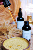 Eine Hand hält eine Pipette mit einer goldbraunen Flüssigkeit, wahrscheinlich Lion's Mane Extrakt, über einer kleinen gelben Keramikschale.