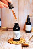 Eine Hand hält eine Pipette mit einem rotbraunen Lion's Mane Flüssigextrakt über der geöffneten Flasche auf einem gewebten Untersetzer.