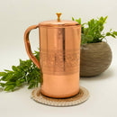 Gravierter Kupfer-Wasserkrug mit Deckel, auf einem runden geflochtenen Untersetzer neben einer grünen Topfpflanze.