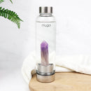 Eine "myga" Wasserflasche mit Amethystkristall, platziert auf einem runden Holzbrett neben einem weißen Tuch und einem grünen Farn auf einer weißen Marmoroberfläche.