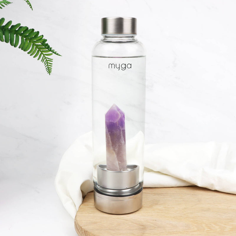 Eine "myga" Wasserflasche mit Amethystkristall, platziert auf einem runden Holzbrett neben einem weißen Tuch und einem grünen Farn auf einer weißen Marmoroberfläche.