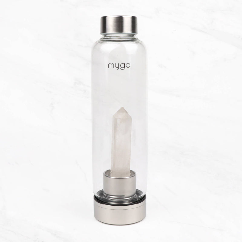 Eine transparente Wasserflasche der Marke "myga" mit einem klaren Quarzkristall im Inneren, stehend auf einer weißen Marmoroberfläche.
