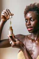 Eine Frau mit strahlender Haut hält eine Flasche Paavani Ayurveda Tridoshic Serum hoch, wobei die Pipette darüber gehalten wird.