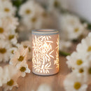 Eine beleuchtete, keramische Duftlampe/Wärmediffusor mit einem ausgeschnittenen Bambusmuster steht auf einer Holzoberfläche, umgeben von unscharfen weißen Gänseblümchen.