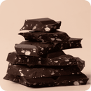 Ein Stapel unregelmäßiger dunkler Schokoladenstücke mit kleinen hellen Nussstückchen darauf, präsentiert vor einem schlichten, beigefarbenen Hintergrund.