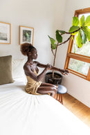 Eine dunkelhäutige Frau mit lockigem braunem Haar in einem goldenen Kleid sitzt auf einem Bett und trägt Körperöl auf ihren linken Arm auf, während sie aus einem großen Holzfenster blickt. Eine Topfpflanze und gerahmte Kunstwerke sind in der Nähe.