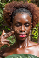 Eine Frau mit leuchtend rosafarbenen Lippen trägt eine Gesichtsmaske auf ihre Wange auf und blickt direkt in die Kamera, umgeben von tropischen Blättern.