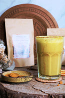 Ein hohes Glas goldgelbe Latte mit Gewürzen, einer kleinen Schale Pulver und Mischungsverpackungen auf einer Holzscheibe.