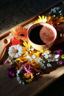 Ein dunkles, schaumiges Getränk in einer gelben Keramiktasse ist von einem lebendigen Arrangement frischer Blumen in Gelb-, Pink-, Weiß- und Lilatönen umgeben, alles auf einem hellen Holztablett. Ein kleiner Holzlöffel liegt neben der Tasse, während ein starker diagonaler Schatten die Szene durchschneidet.