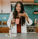 Eine Frau mit langen, grau melierten Haaren steht in einer Küche, hält eine Tasse Tee und riecht daran; eine Dose Woman's Blend Tee steht auf der Theke.