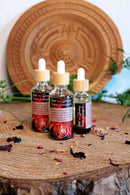 Drei Tropfflaschen "Hot n Spicy Tantric Massage Oil" sind auf einem runden Baumstumpf aus Holz arrangiert. Getrocknete rote und schwarze Blütenblätter sind um die Flaschen verstreut, und grünes Laub sowie eine geschnitzte Holzplakette sind im Hintergrund sichtbar.