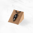 Ein glänzender, geschnitzter schwarzer Obsidian-Pfeilspitzen-Anhänger an einer schwarzen Kordel, präsentiert auf einem kleinen Schmuckständer aus Pappe vor weißem Marmorhintergrund.