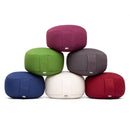 Stapel von sechs runden Meditationskissen in Uni-Farben: Burgunderrot, Grün, Grau, Blau, Weiß und Rot, mit plissierten Seiten und Logo-Etiketten, auf weißem Hintergrund.