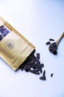 Ein geöffneter Kraftpapierbeutel von „Ayamanu Naturals“ mit rohen Schokoladenstücken entleert sich auf einen weißen Hintergrund. Ein Holzlöffel, ebenfalls mit Schokoladenstücken gefüllt, liegt neben den verstreuten Stücken und betont die biologische und natürliche Beschaffenheit des Produkts.